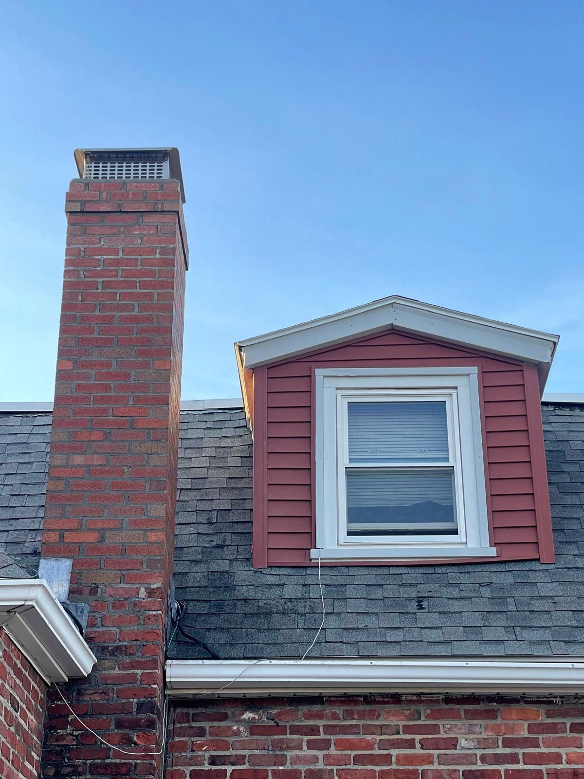 A brick chimney standing next to a house roof with a pink-sided dormer window against a clear blue sky.