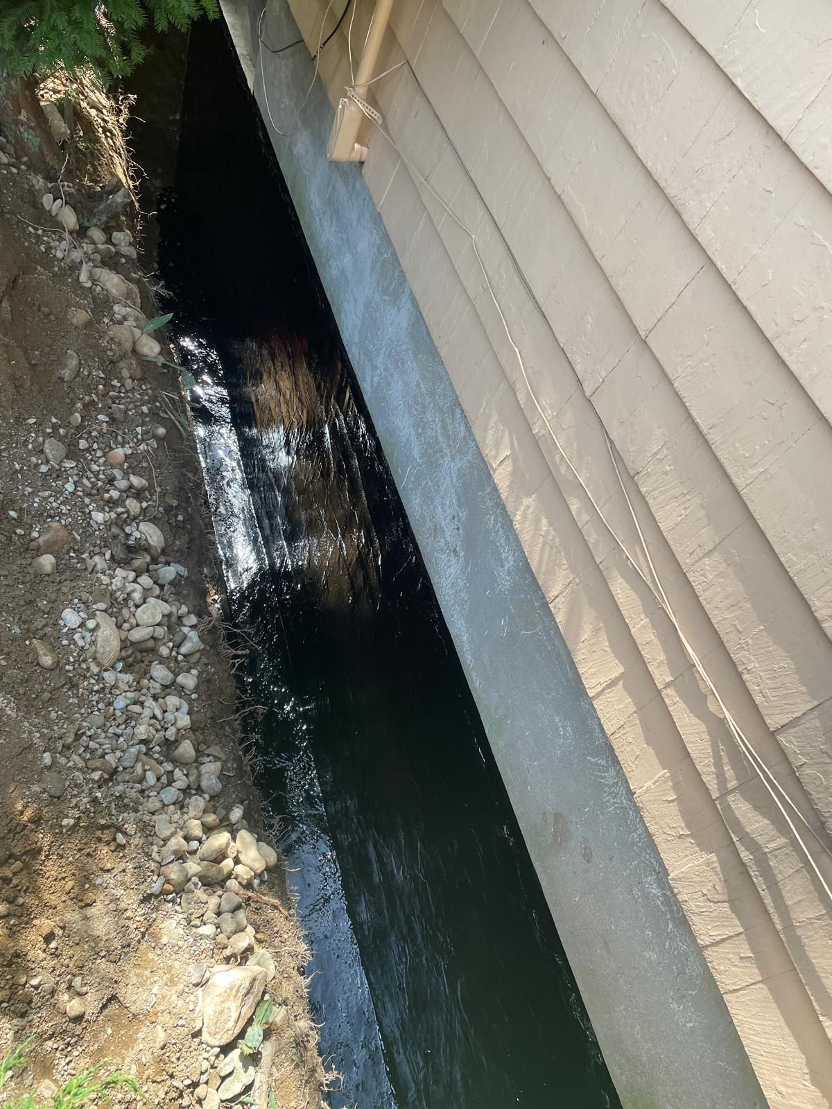 A trench dug alongside a house foundation, showing a black waterproofing membrane applied to the concrete wall.