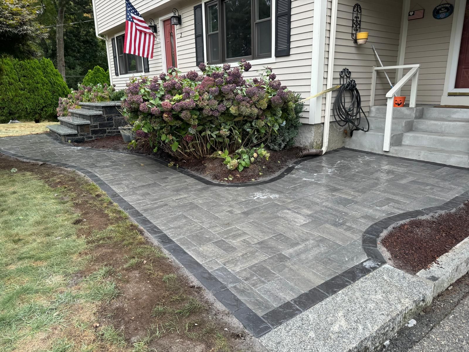 A gray stone patio and walkway lead to a house entrance with concrete steps and a large flowering bush.