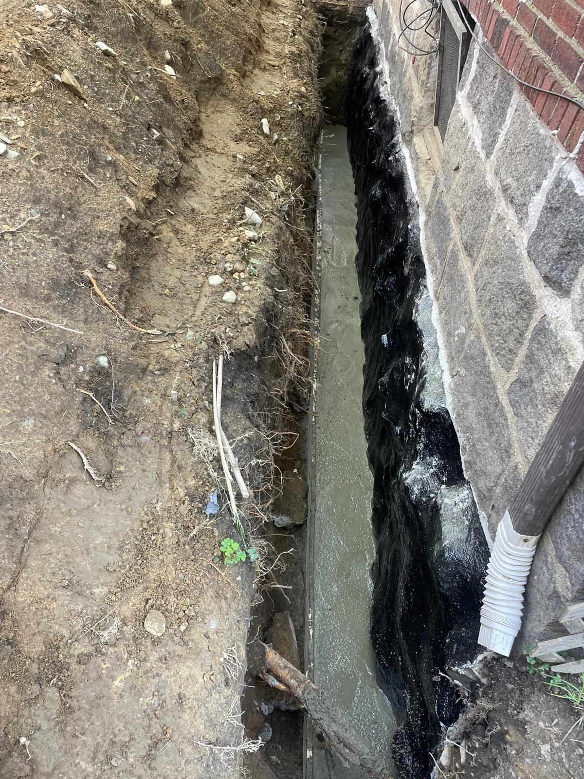 A vertical trench beside a brick foundation wall treated with black waterproof coating and a fresh concrete footing.