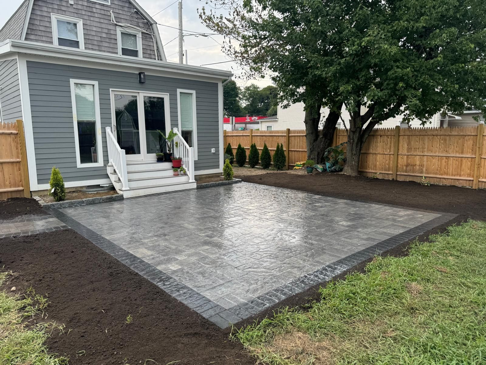 A gray house with white stairs leading to a newly installed rectangular stone patio surrounded by dark soil and grass.