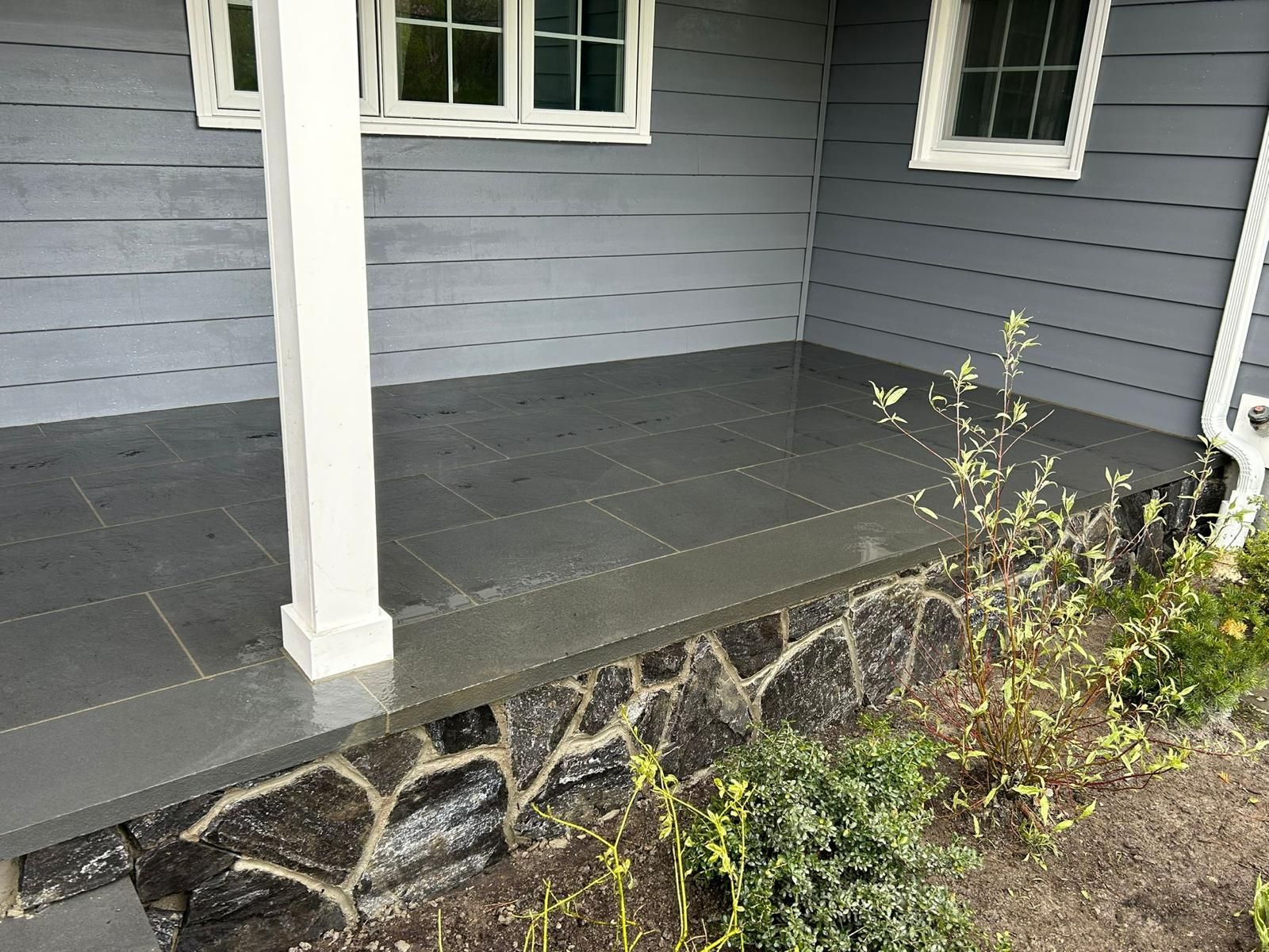 A porch with gray tiled flooring, a white pillar, gray horizontal siding, and a stone-faced foundation.