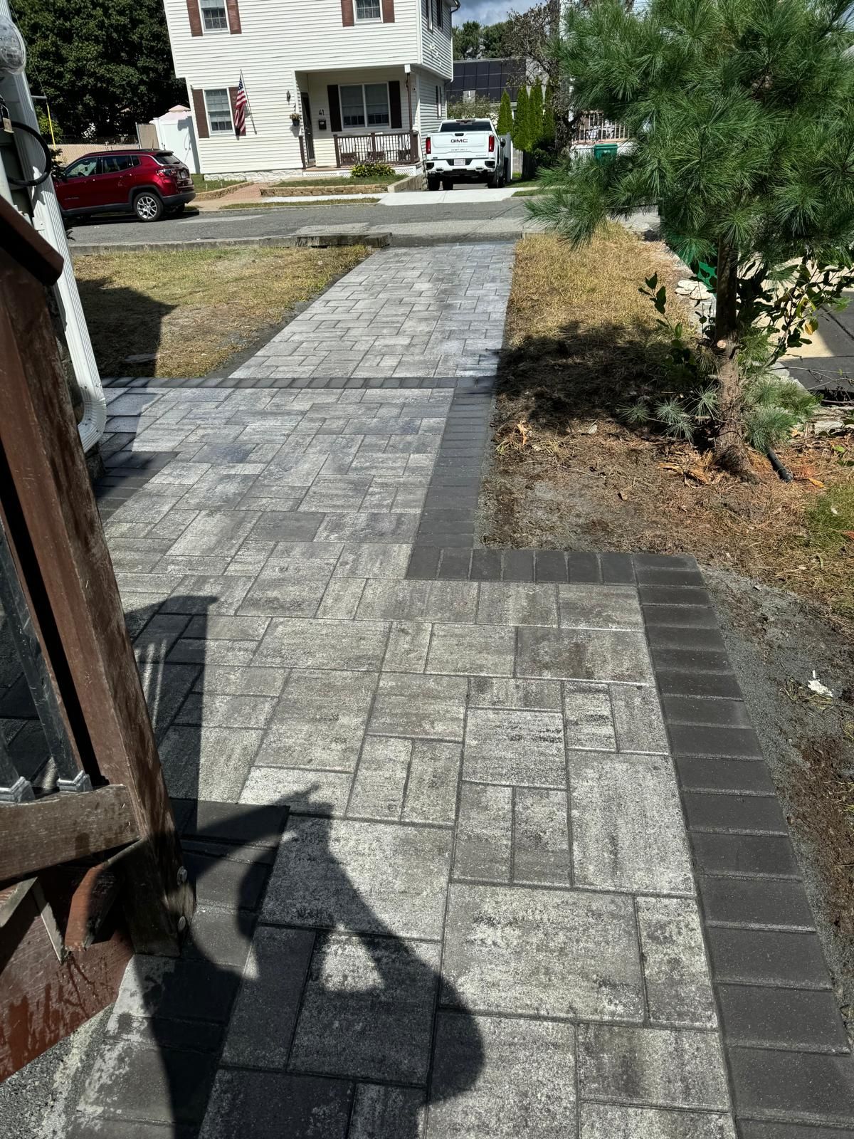 A residential stone paver walkway leads toward a street, featuring a grey patterned main area with a dark border.