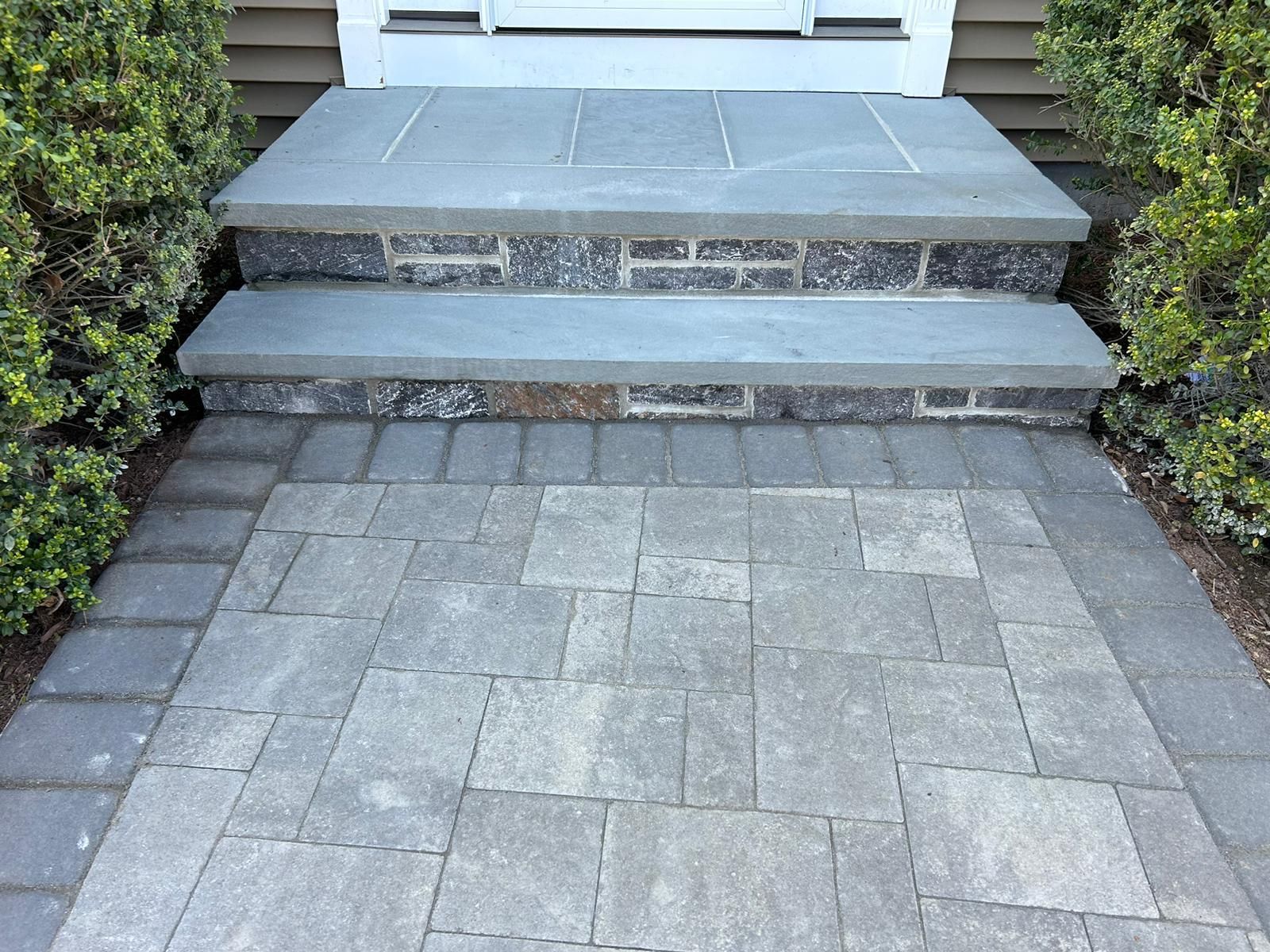 Two stone steps leading to a doorway, centered above a patio made of grey rectangular pavers with a border.