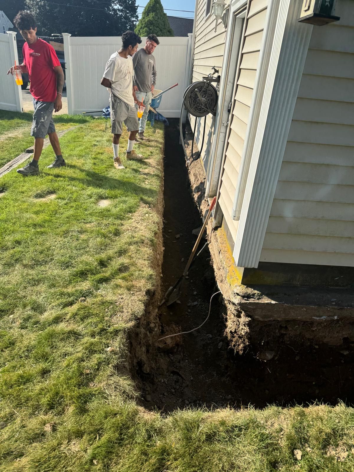 Three people stand near a deep trench dug alongside the foundation of a house in a residential yard.