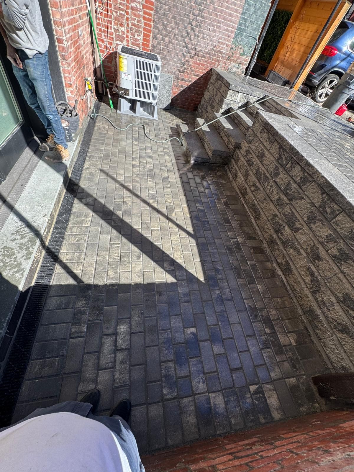 A newly installed brick-paver patio features a light-colored central area bordered by dark-toned bricks next to a wall.