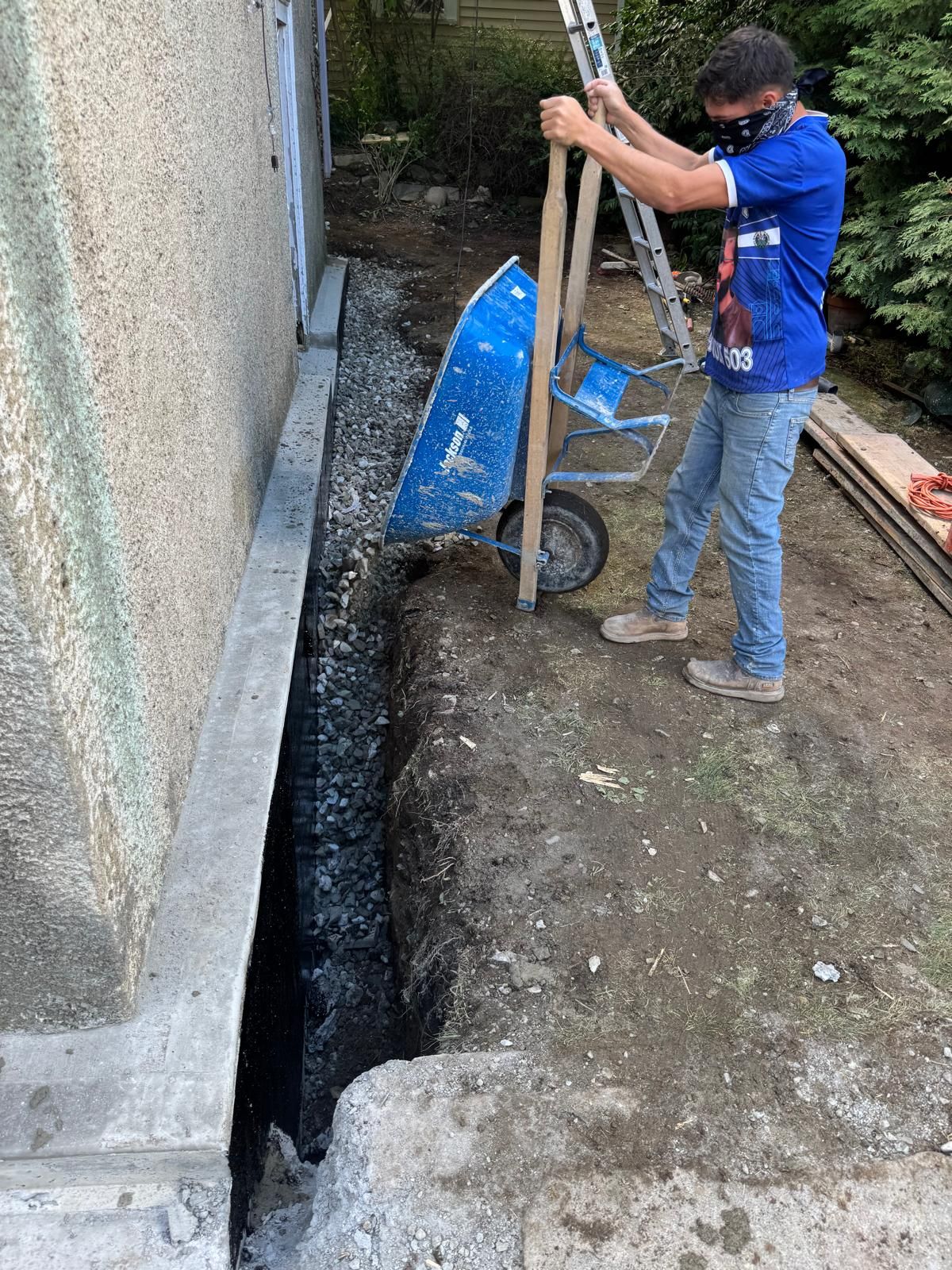 A person in a blue shirt holds a blue wheelbarrow near a narrow trench being dug alongside a building’s foundation.