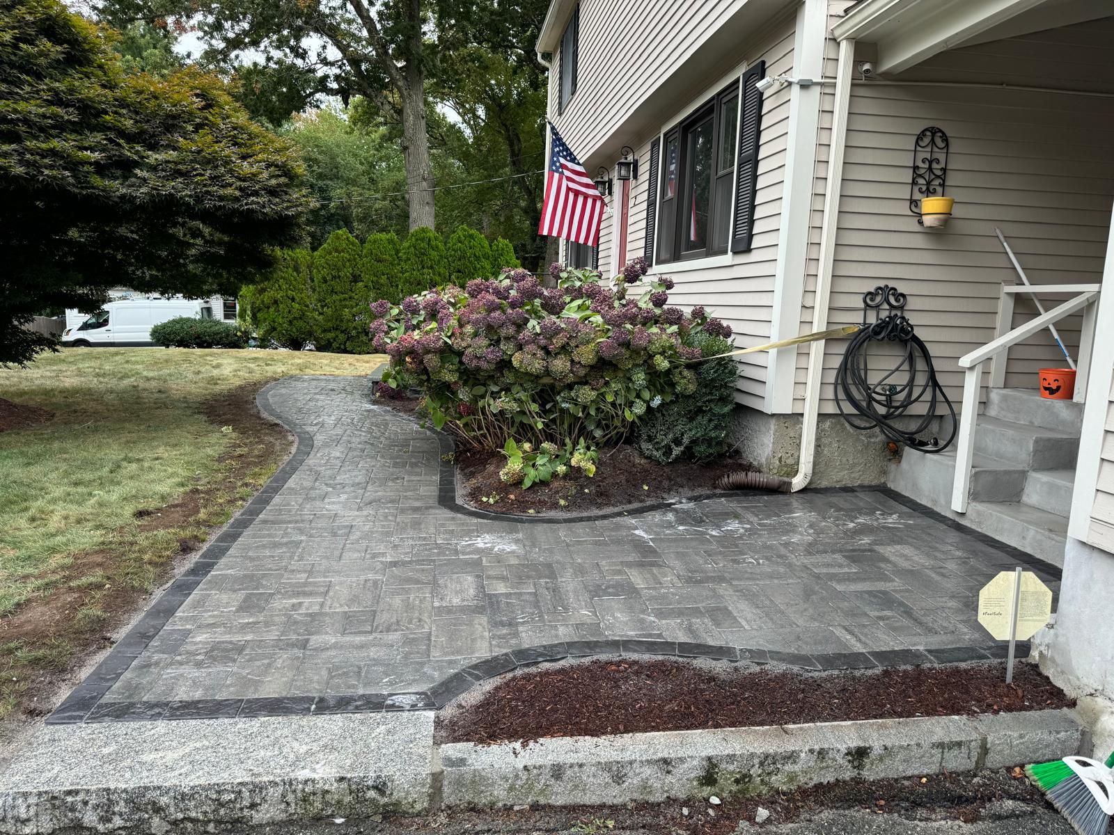 Newly installed paver walkway and patio leading to a house entrance with landscaping, a US flag, and stone borders.