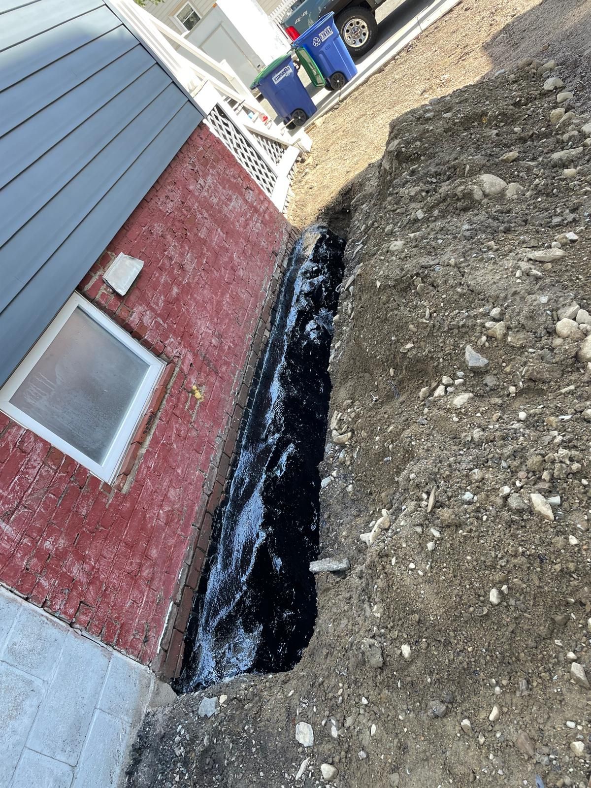 A trench dug along the side of a red brick house foundation, coated with black sealant, next to a pile of loose dirt.