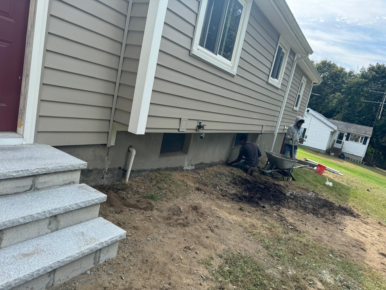 Two workers landscape the soil beside a beige house with gray steps and a maroon door.