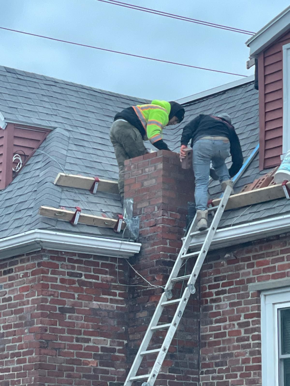 Two construction workers in high-visibility gear work on a residential brick chimney on a shingled roof with roof jacks.