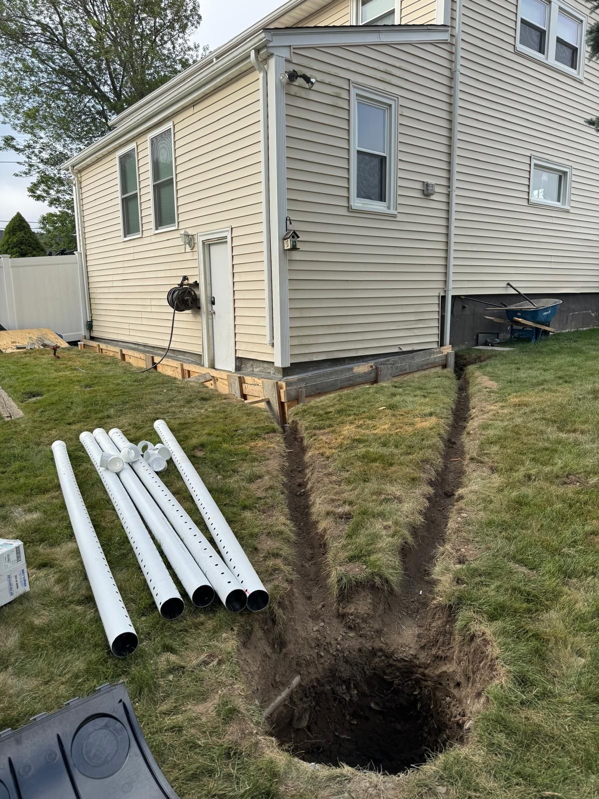 A side view of a house with siding, showing a trench dug in the lawn leading to a hole near PVC drainage pipes.