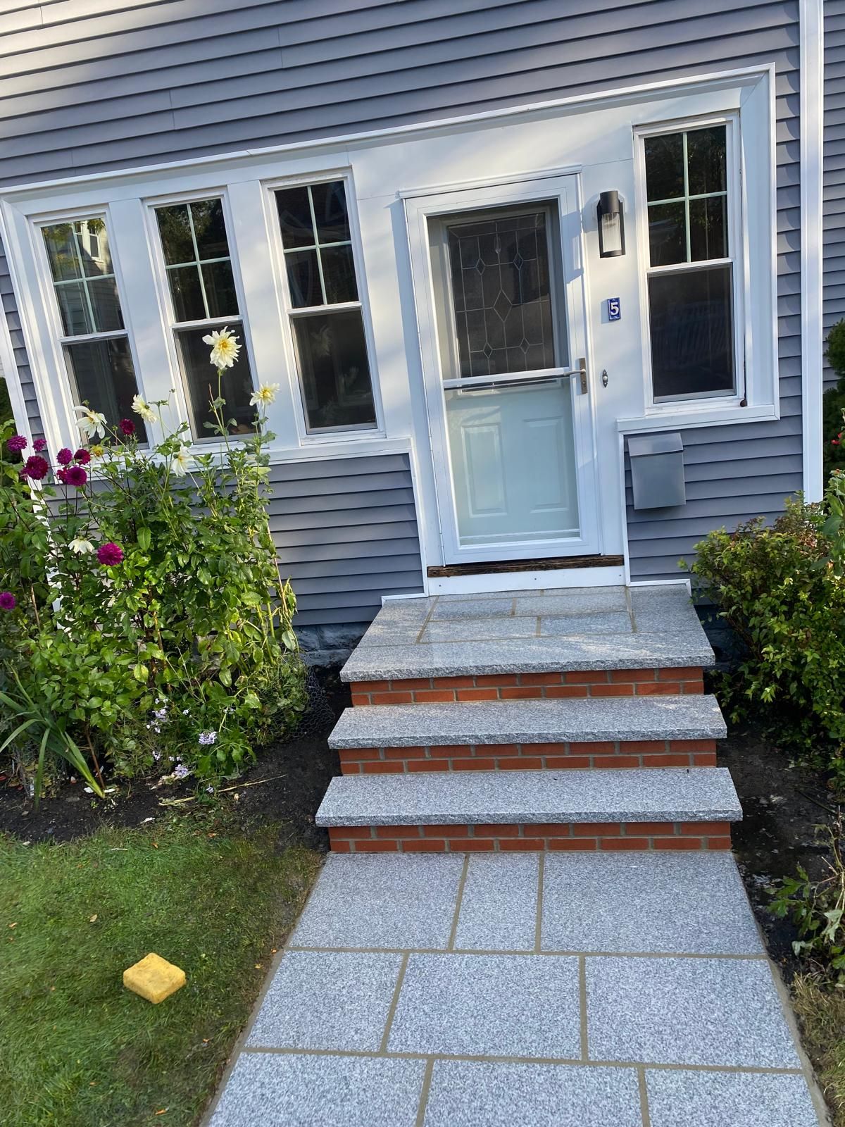 A gray house entrance with stone steps leading to a light blue front door, flanked by windows and landscaped greenery.