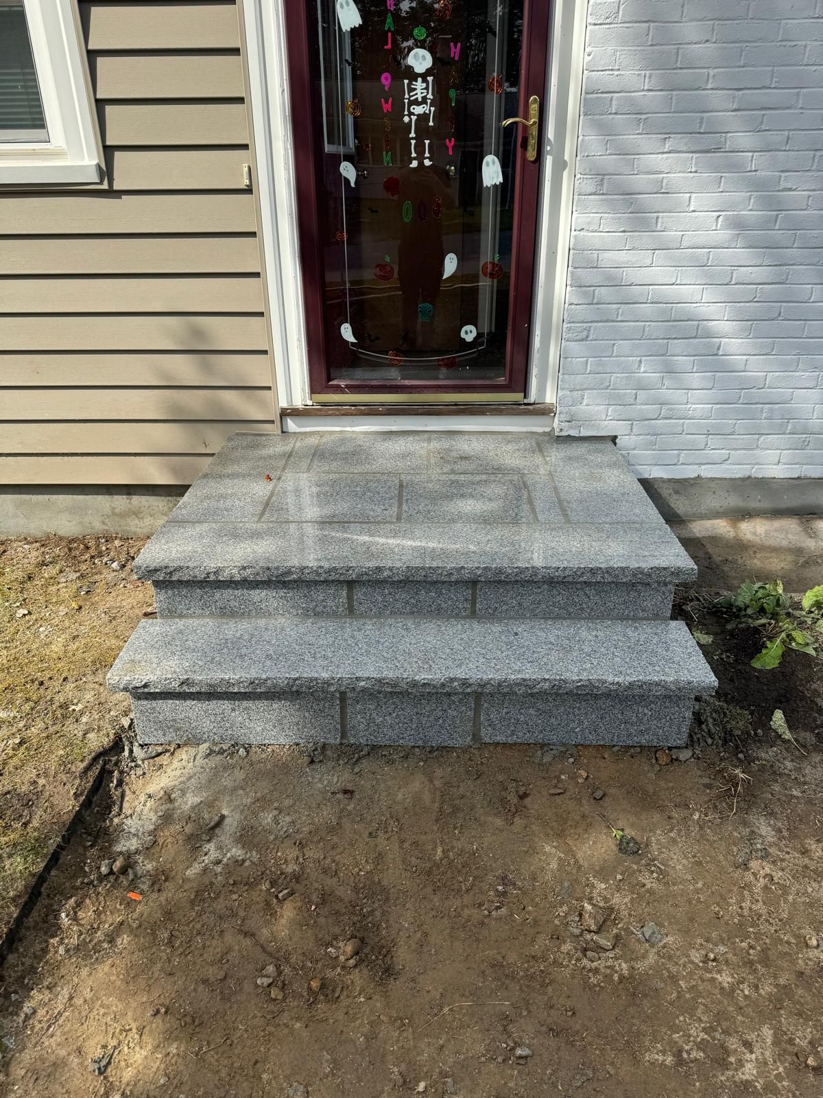 Two new gray speckled granite steps leading up to a front door with a burgundy storm door and white brick siding.