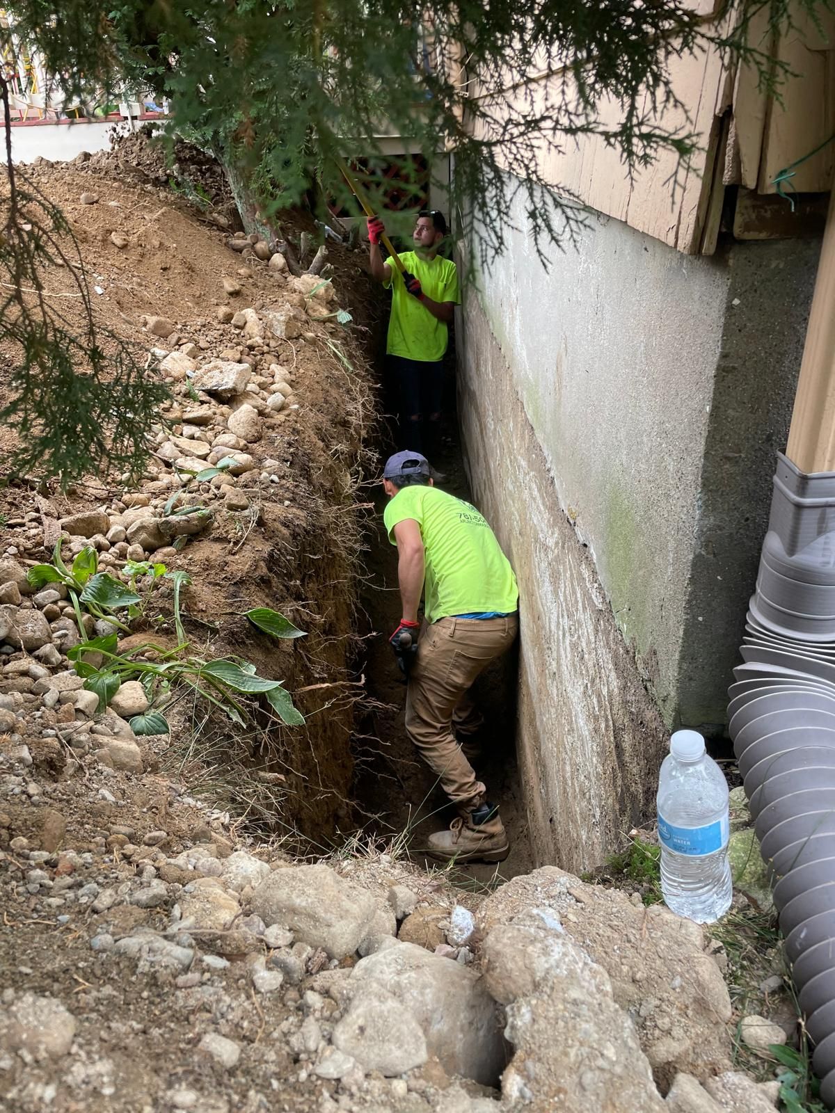 Two people in high-visibility shirts dig a narrow trench along the foundation of a building beside a pine tree.
