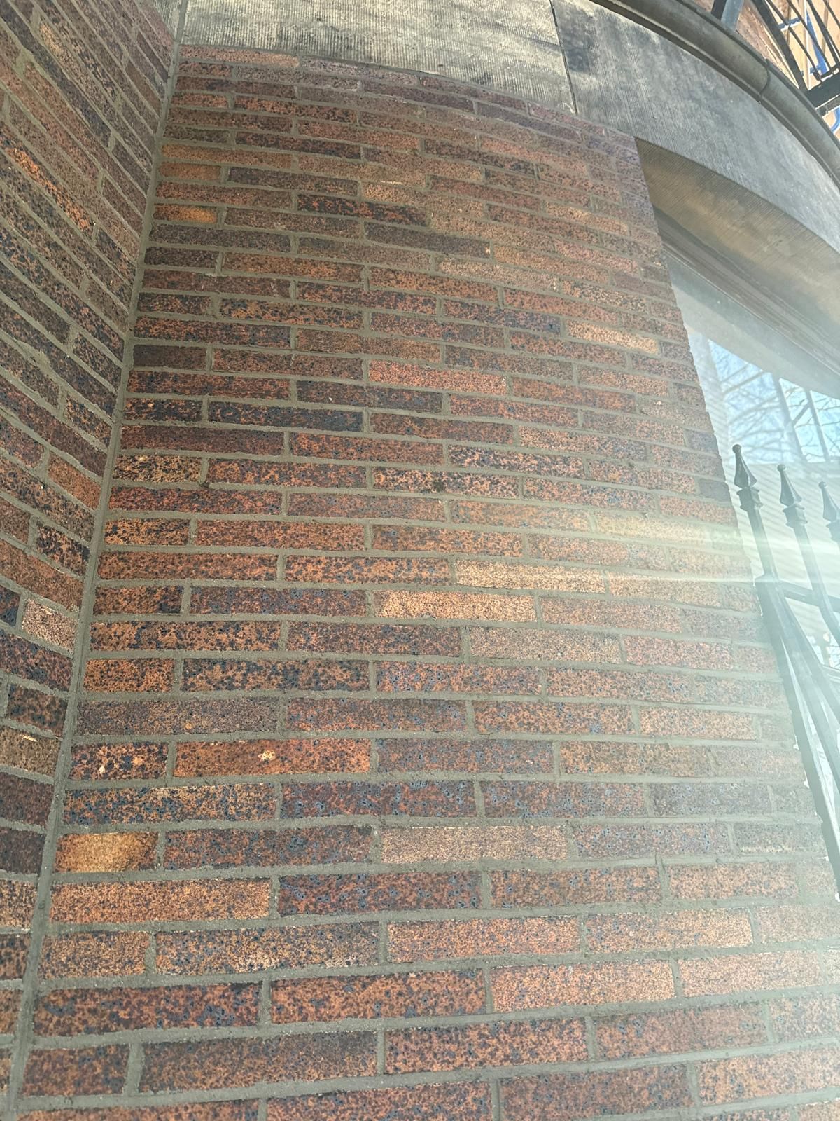 An angled view of an exterior wall made of textured, dark brown bricks, with a bright sun flare across the right side.