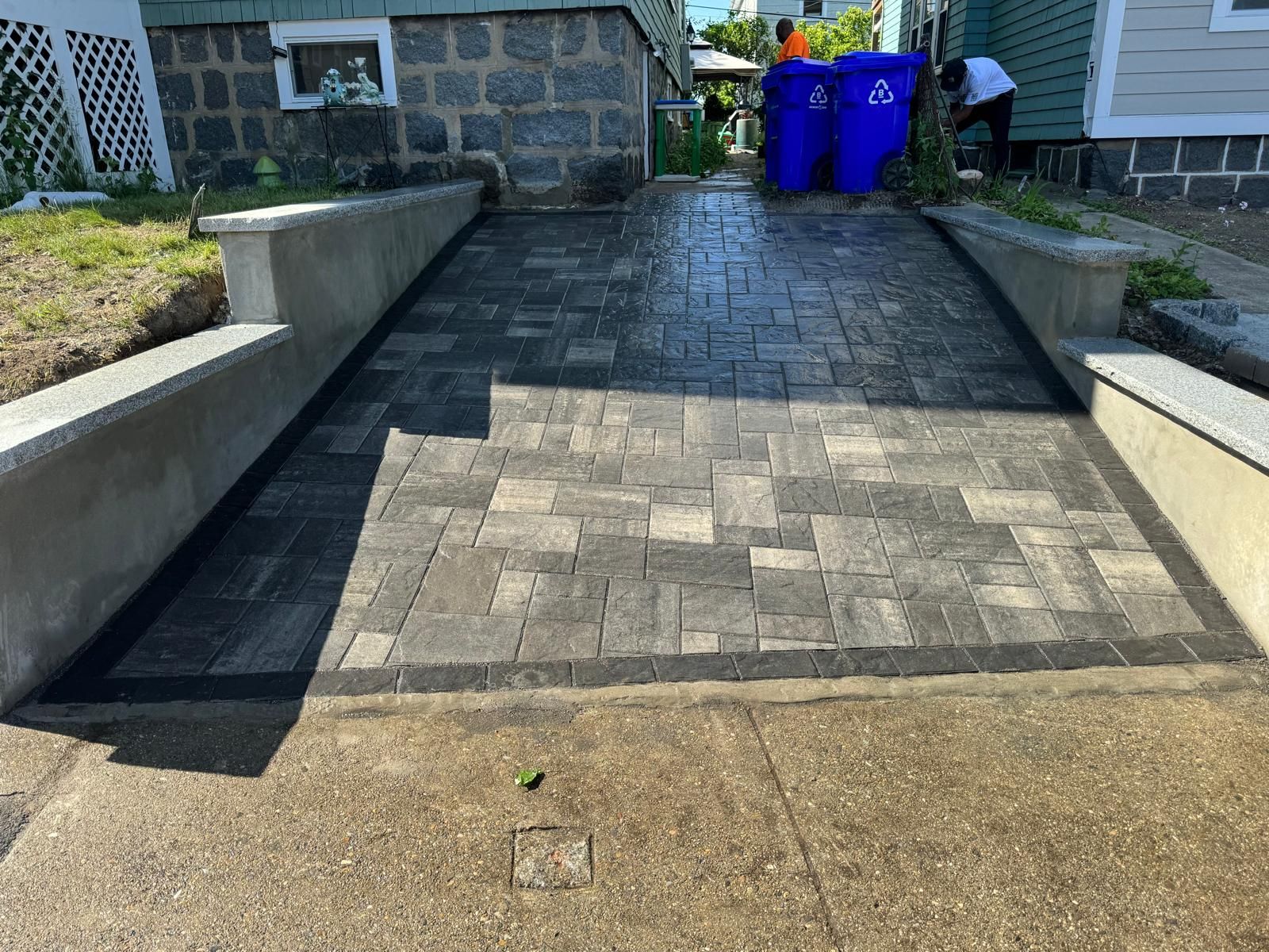 A new paver driveway leads to a stone house foundation, flanked by concrete retaining walls and blue recycling bins.