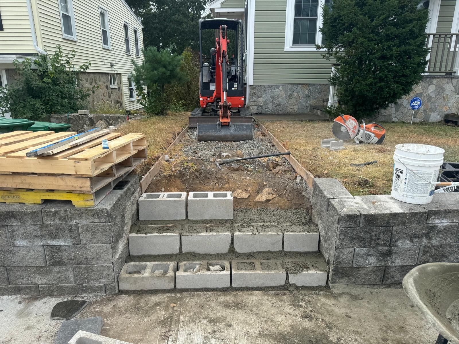 A small excavator sits in the yard of a house during a masonry construction project with steps made of cinder blocks.