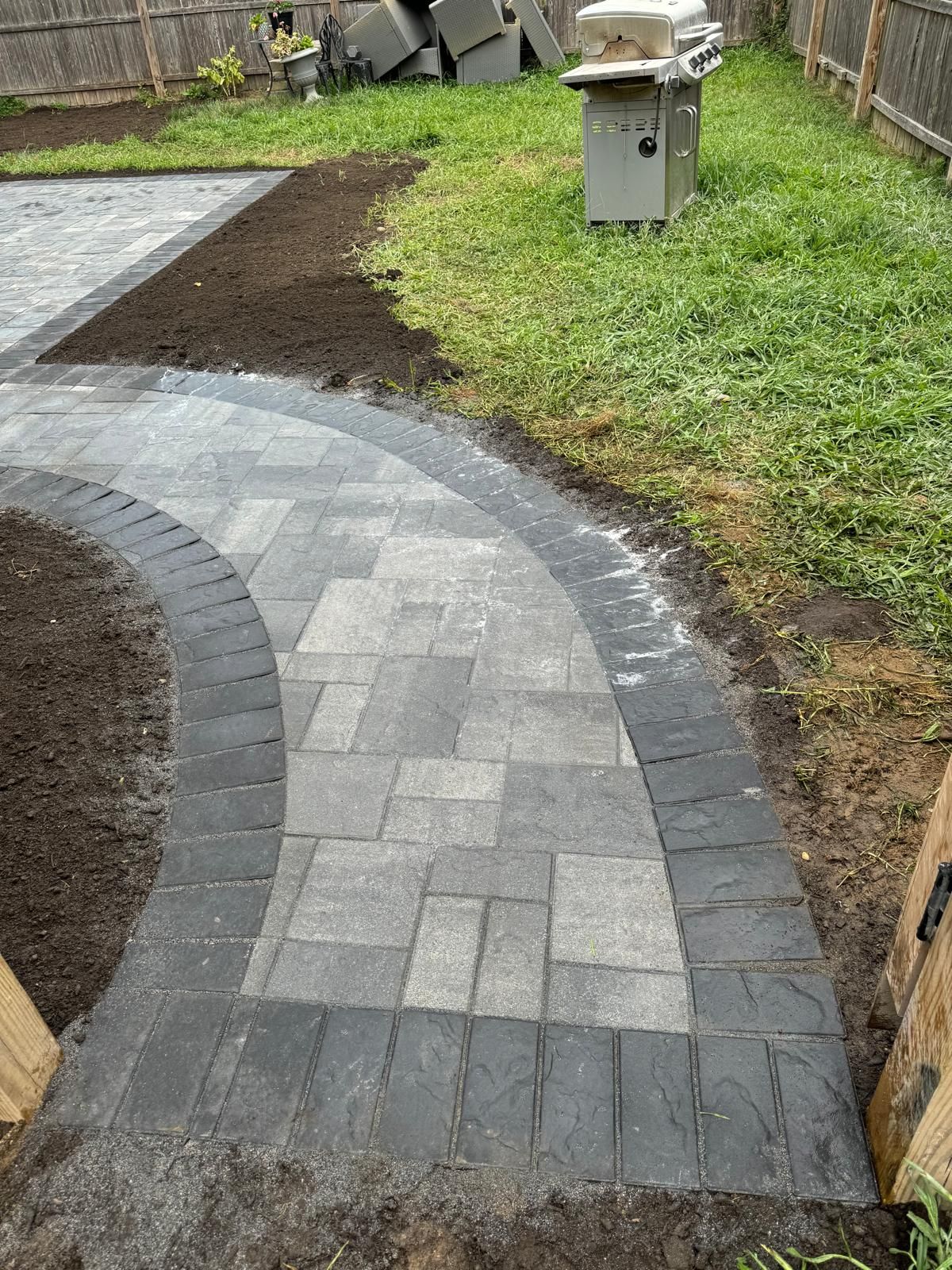 A gray stone walkway curves through a backyard, bordered by dark charcoal bricks next to sections of fresh soil and lawn.
