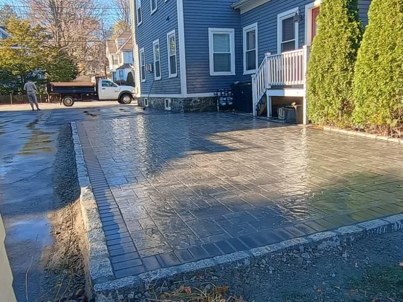A newly installed grey paver patio beside a blue house with a person standing near a pickup truck in the background.