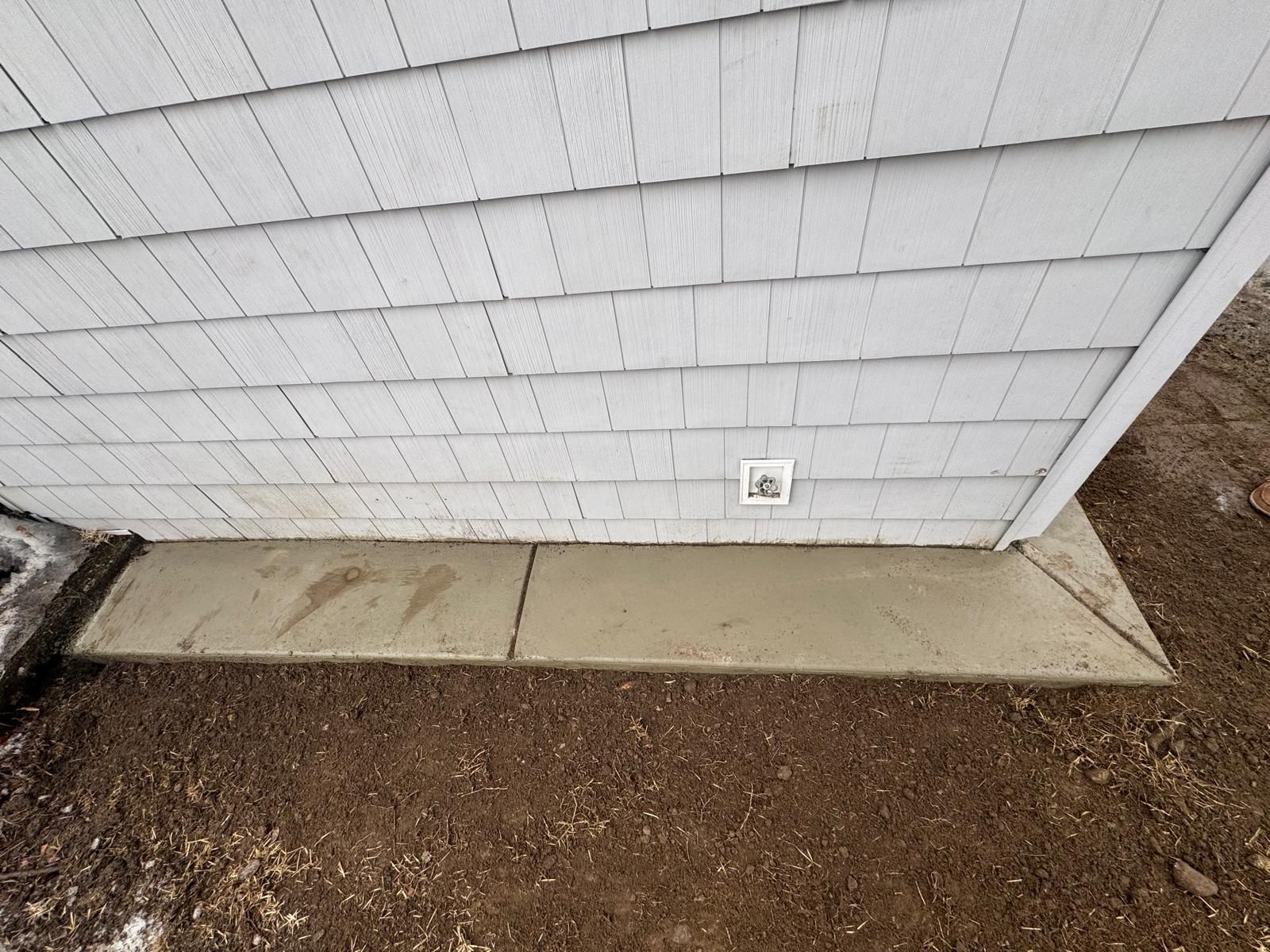 A newly poured concrete walkway along the base of a light gray, shingled exterior wall with an electrical outlet.
