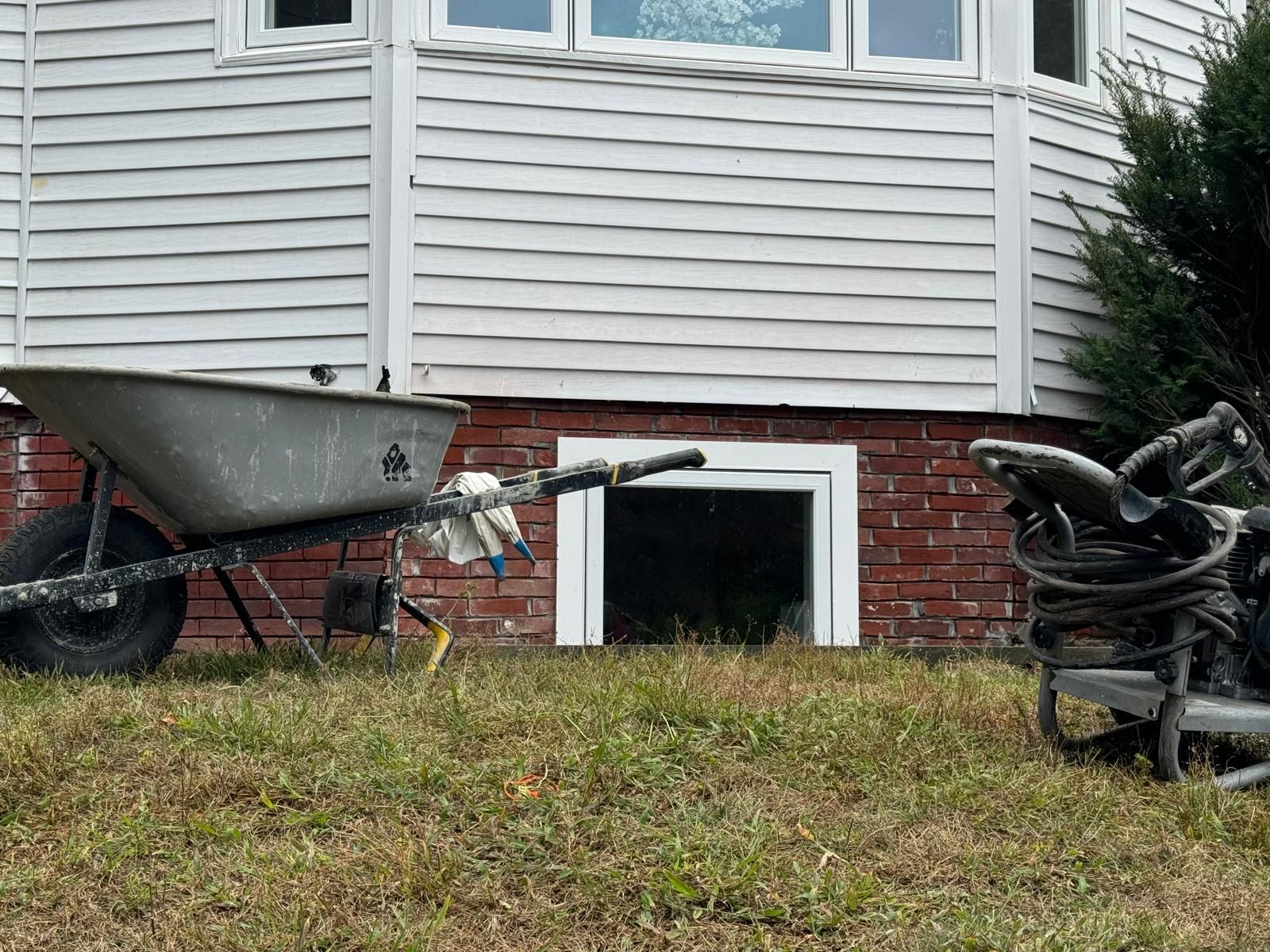 A lawn featuring a wheelbarrow and a piece of equipment sitting in front of a house with white siding and a brick foundation.