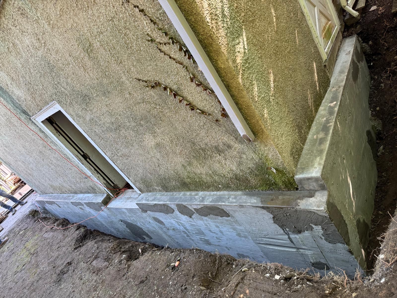 A fresh, grey concrete foundation wall has been poured along the exterior of a textured, mossy-colored building.