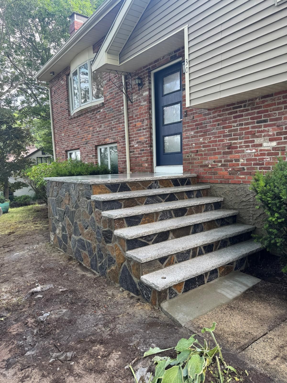 A house entrance with a modern dark blue front door and new stone-clad steps leading up to a brick and siding facade.