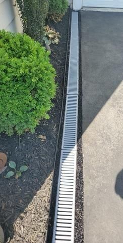 A metal trench drain runs between a paved driveway and a garden bed filled with dark mulch and a green bush.
