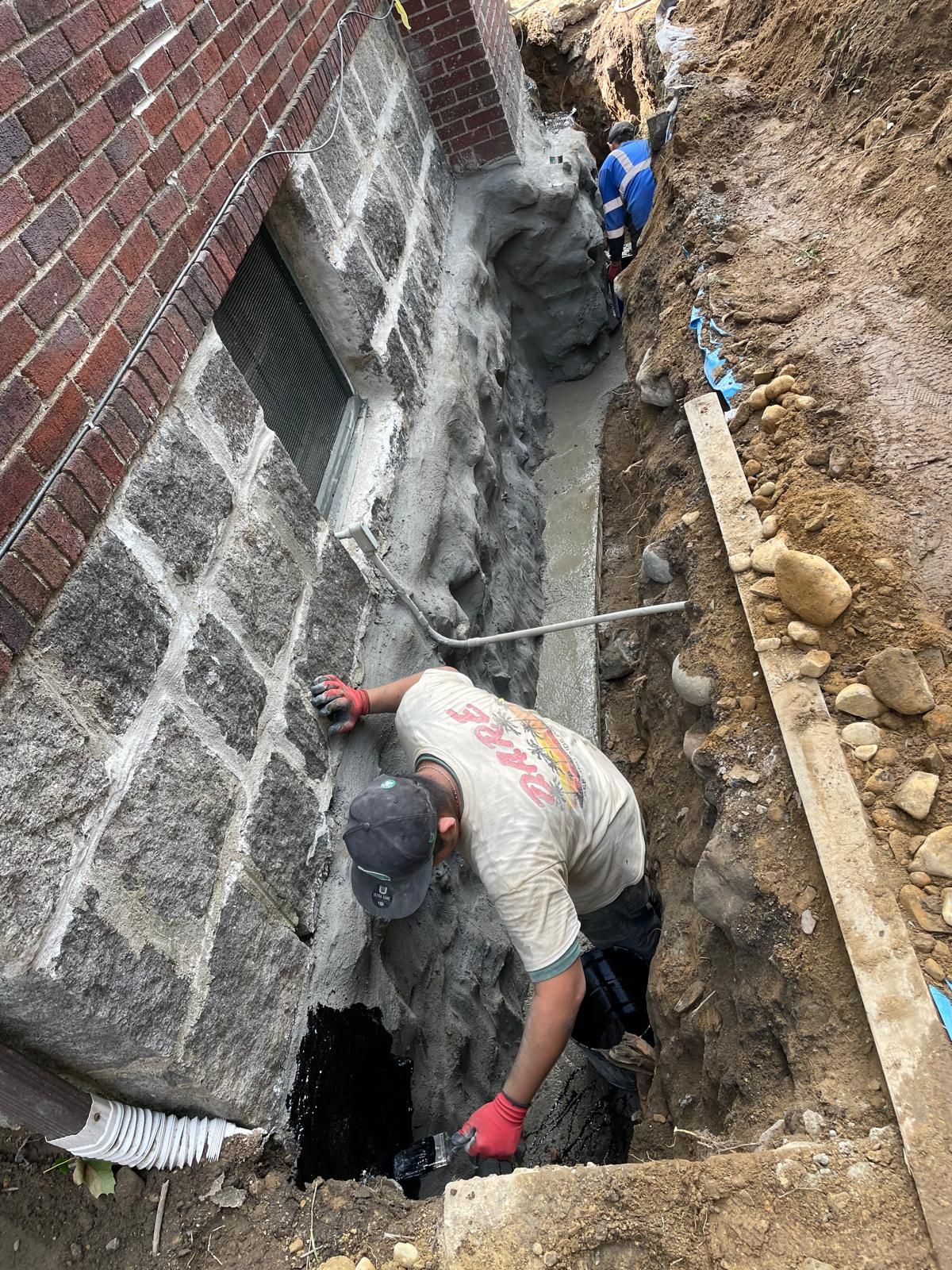 A worker in a trench repairs a stone foundation wall beside a brick house, applying wet concrete to the exterior.