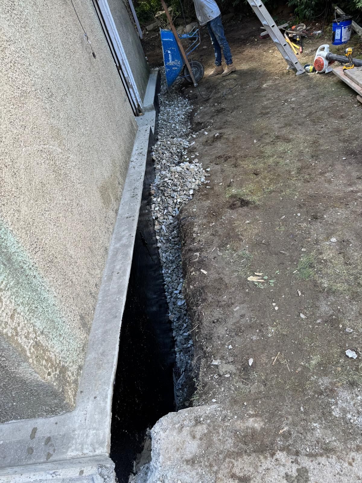 A trench along a concrete foundation, partially filled with gravel, alongside a person standing in a grassy yard.