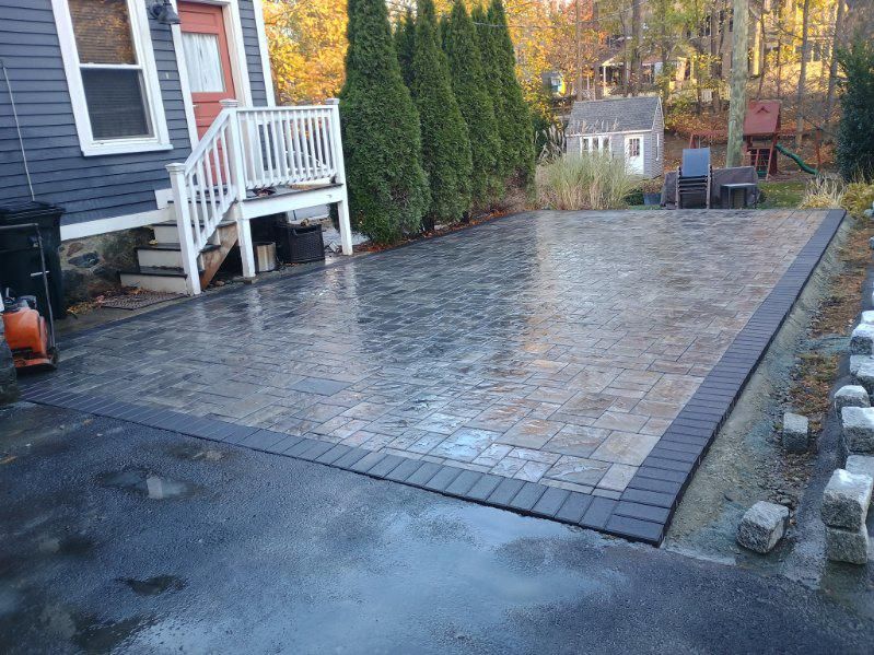 Newly installed stone paver patio with a dark border next to a gray house and asphalt driveway.
