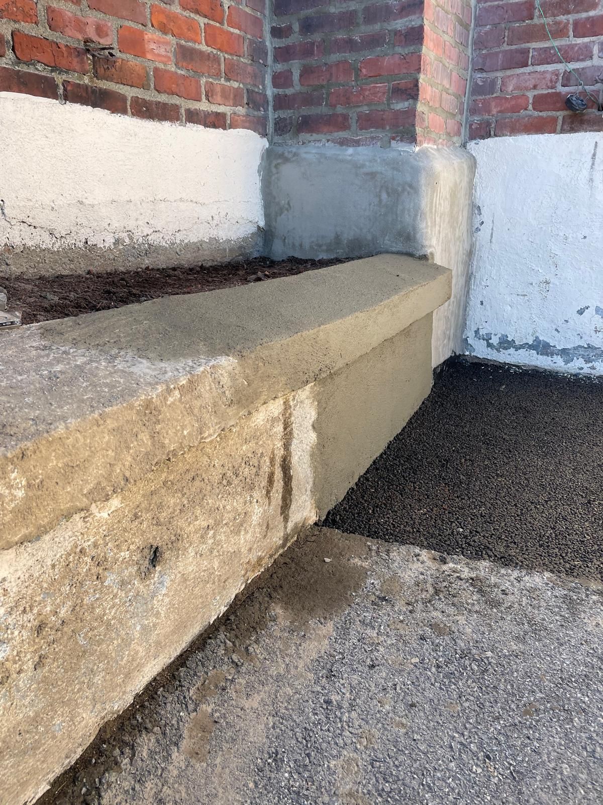 A concrete planter ledge sits against a brick and white-painted wall, with a layer of dark soil and a gravel path nearby.