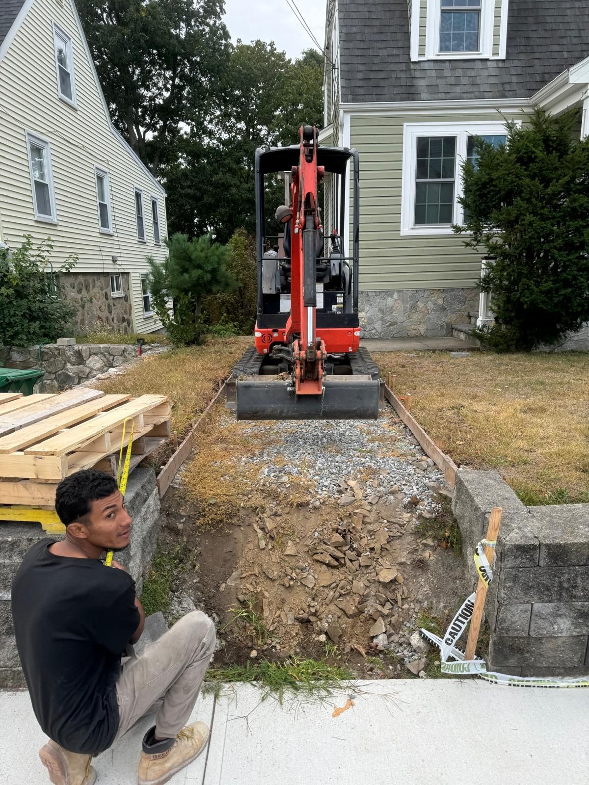 A person crouches near a red mini excavator parked on a gravel walkway leading to a house.