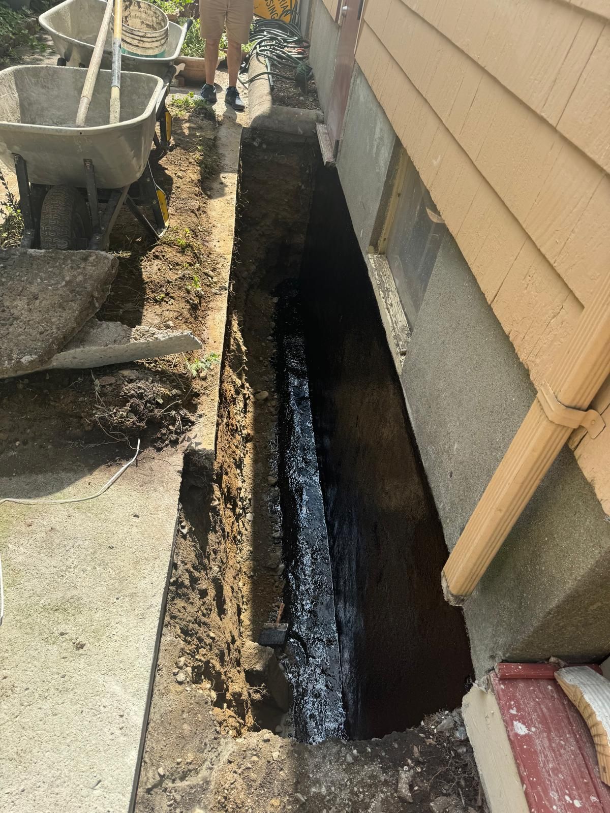 A trench dug along the foundation of a house, revealing a black-coated basement wall next to construction equipment.