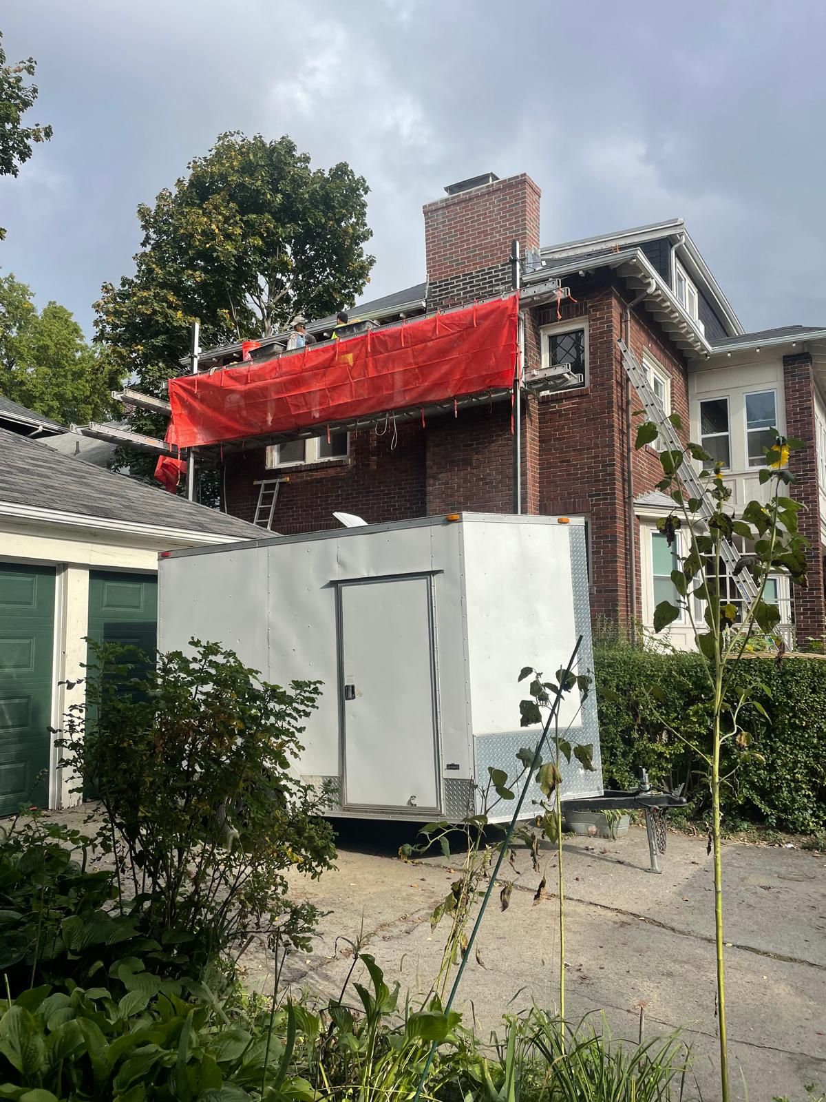A brick house under renovation with scaffolding and a red tarp, featuring a white trailer parked in the foreground.