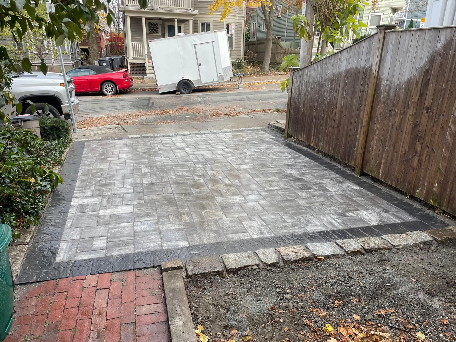 A newly installed stone paver patio sits adjacent to a wooden fence and a red brick walkway in a residential yard.