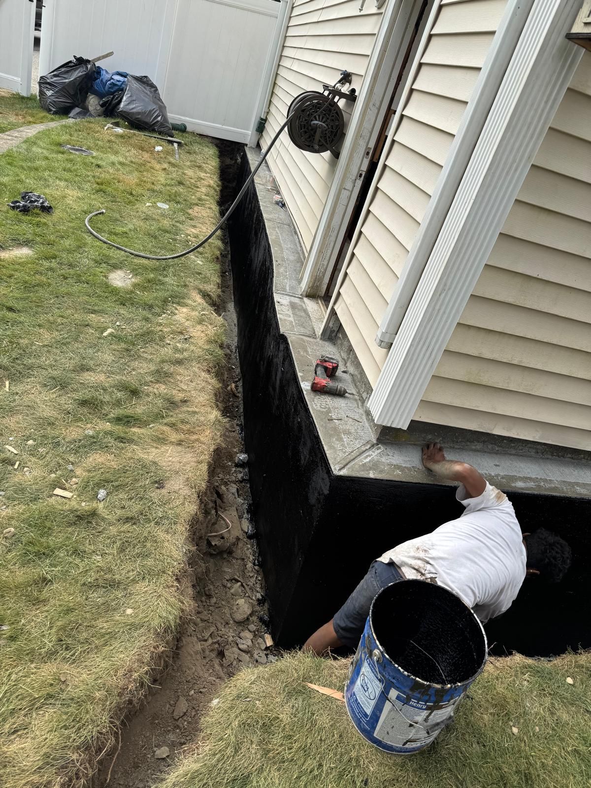 A worker applies black waterproof sealant to a house foundation in a deep trench along the side of a building.