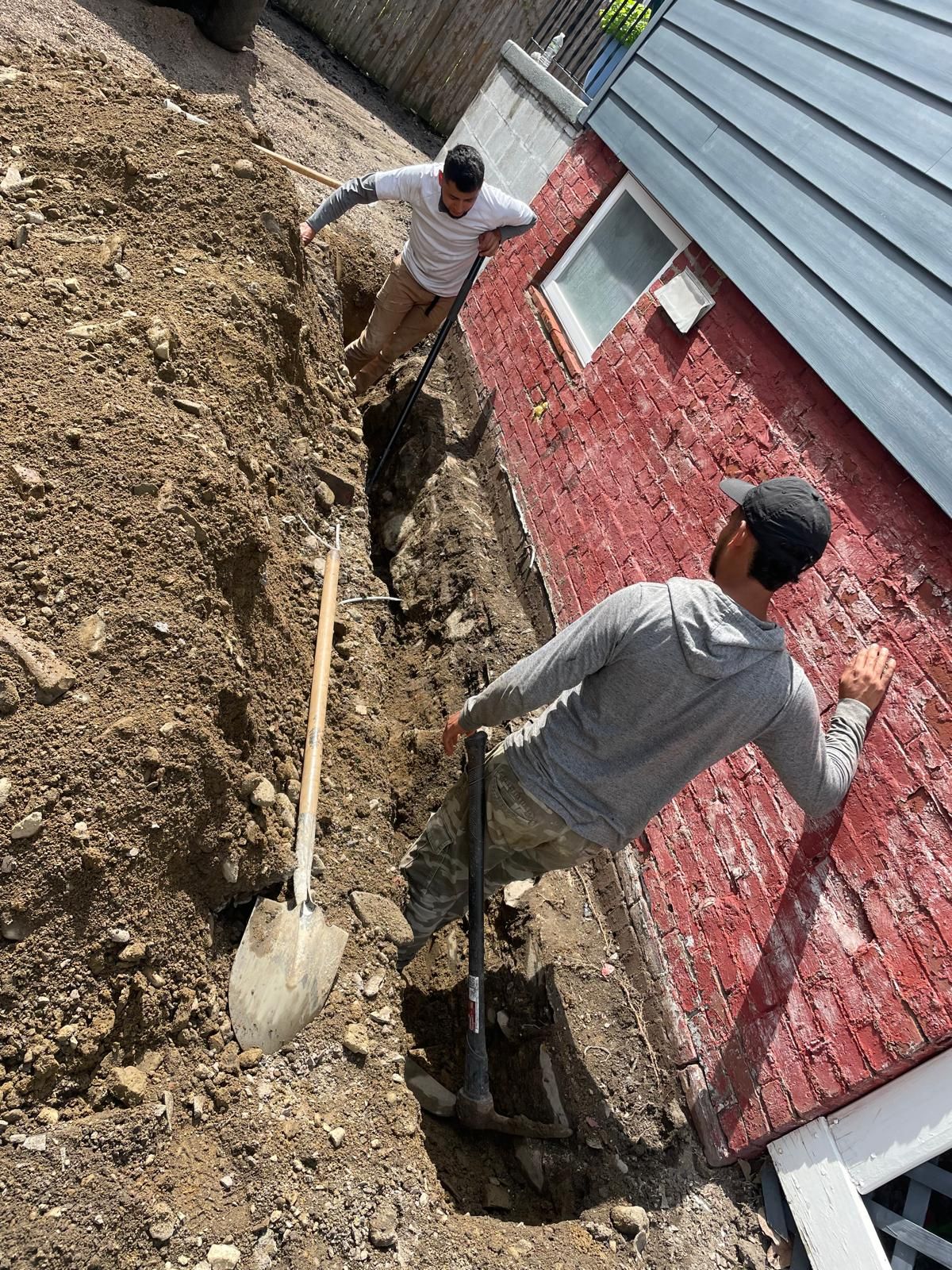 Two people work in a narrow trench next to a red brick foundation wall, using shovels to excavate the earth.