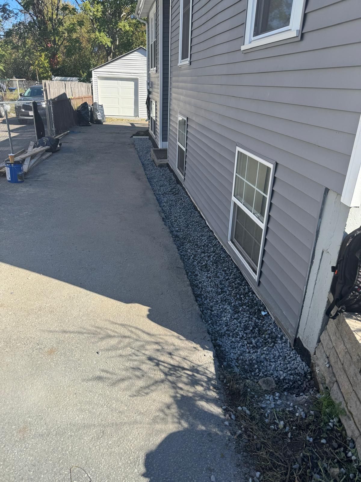A gray-sided house exterior with windows, alongside a paved driveway and a narrow strip of gray gravel at the foundation.