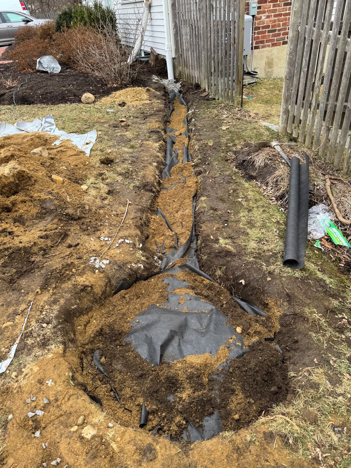 A trench containing black landscaping fabric and drainage pipe in a yard under construction next to a wooden fence.