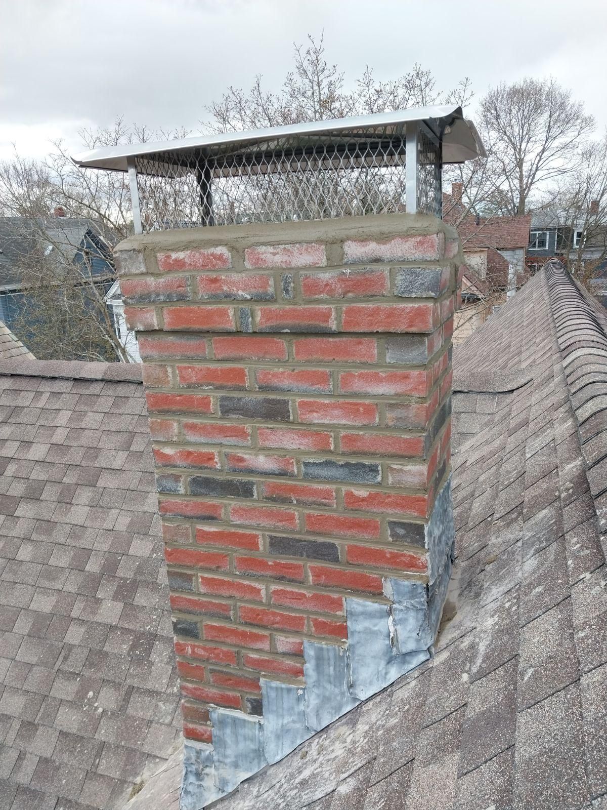 A red brick chimney with metal flashing and a mesh-screened chimney cap, set on a shingled residential roof.