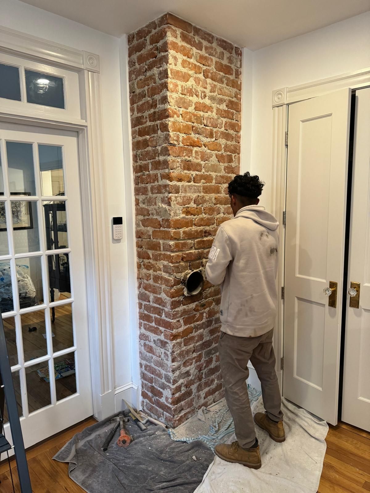 A person in a light hoodie works on a brick chimney column in a room with white doors and hardwood floors.