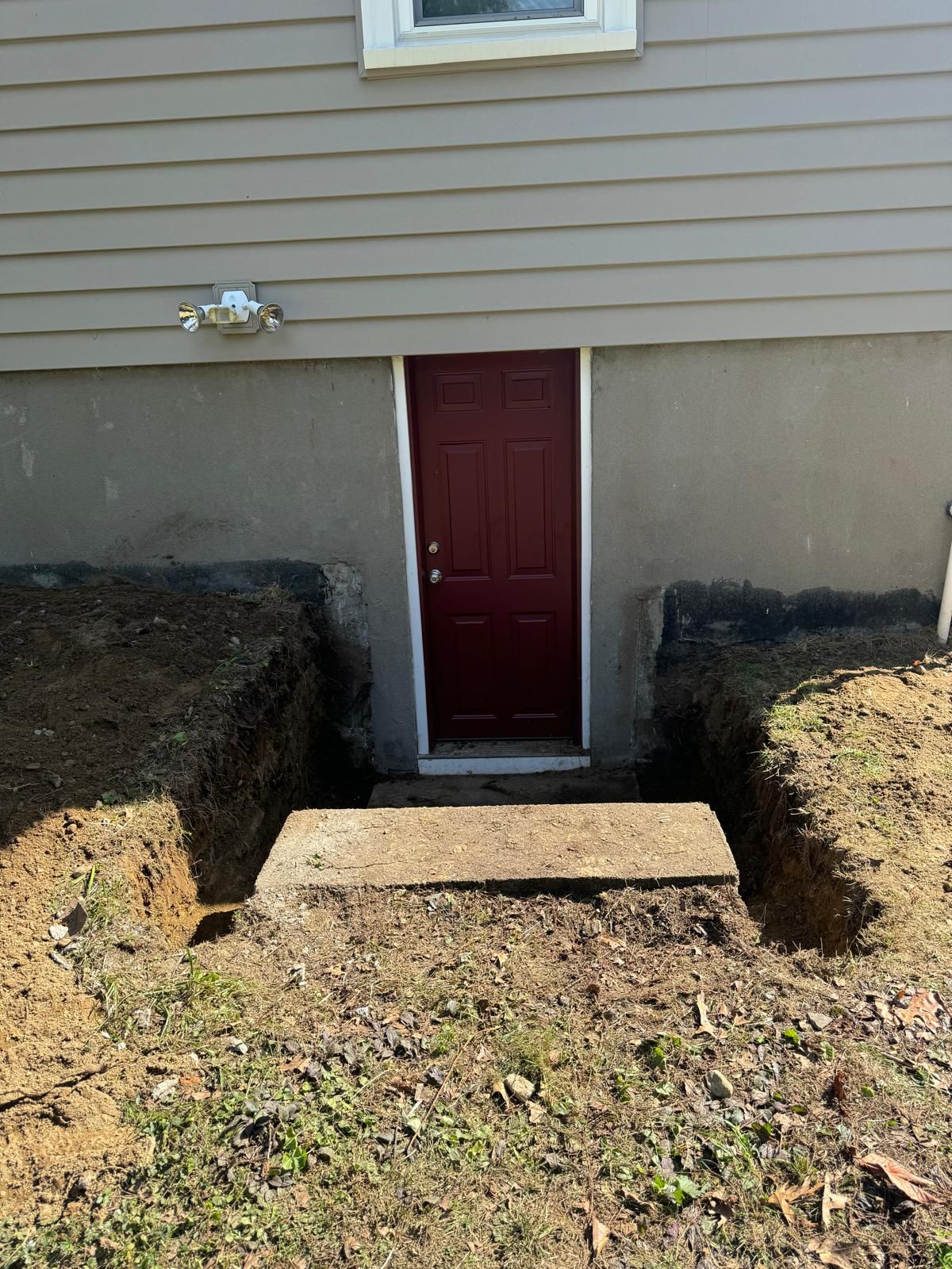 A red exterior door set into a basement foundation, surrounded by a narrow dirt trench and a single concrete step.