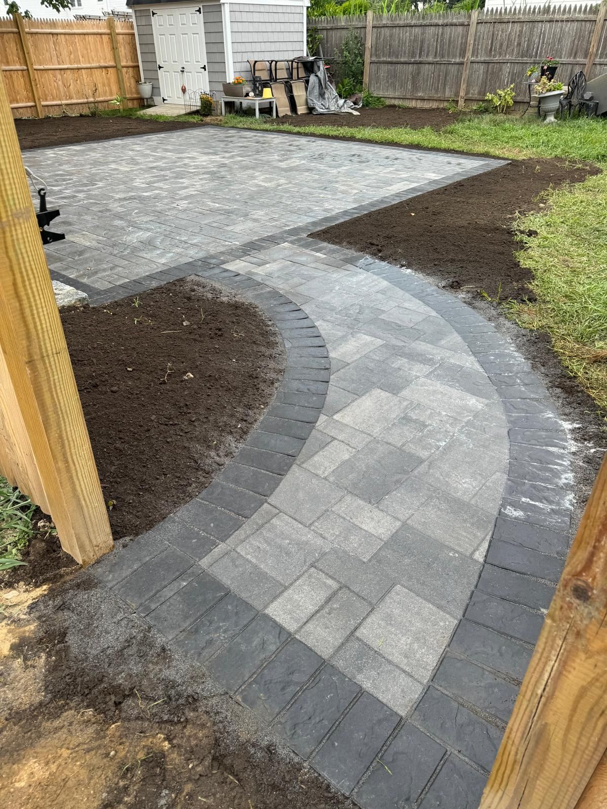 A curved stone paver walkway leading to a larger rectangular patio in a residential backyard with soil and a fence.