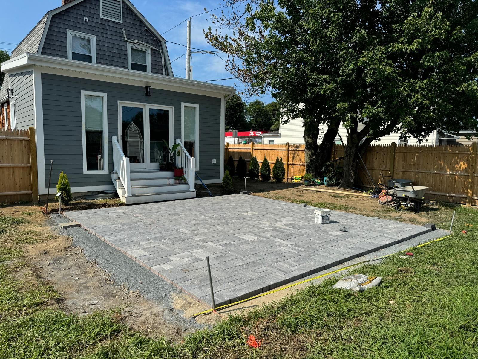 A newly installed grey paver patio in a residential backyard with a grey house, wooden fences, and a large tree.
