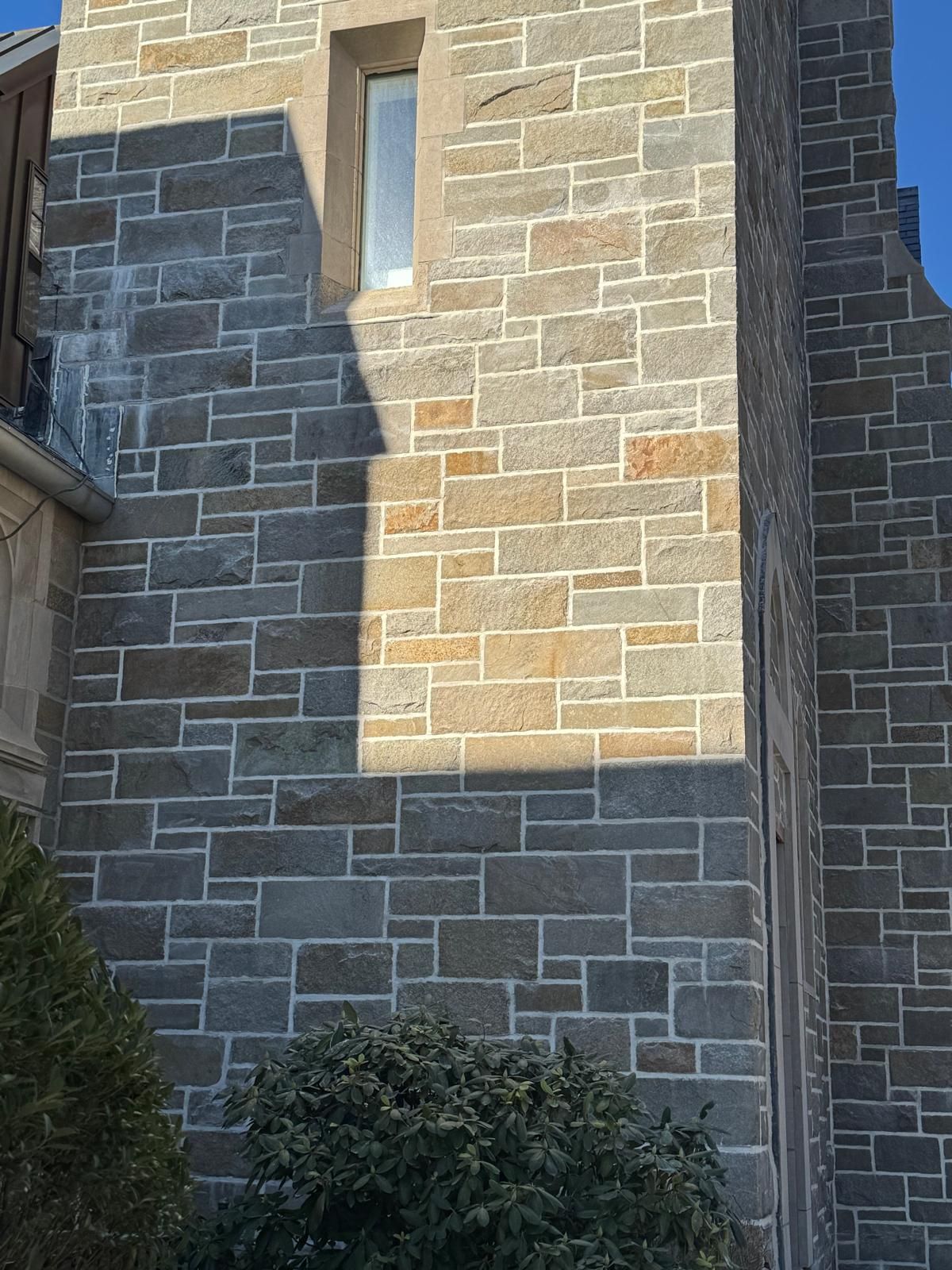 A stone tower wall made of rectangular blocks in shades of gray and tan, with a narrow, vertical window at the top.