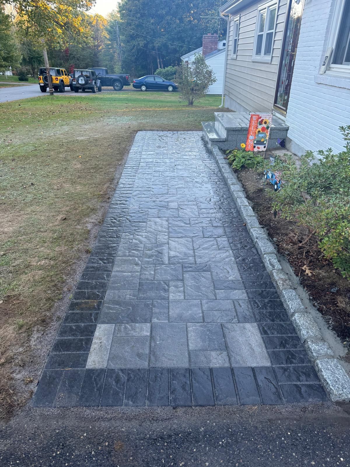 A grey and black paver walkway leads from an asphalt driveway to the front steps of a beige house.