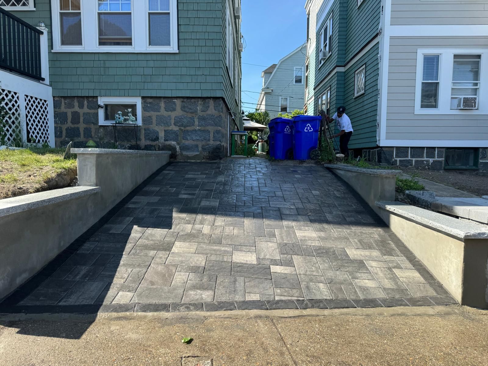 A paved driveway with a rectangular stone pattern leads between two houses, framed by concrete retaining walls.