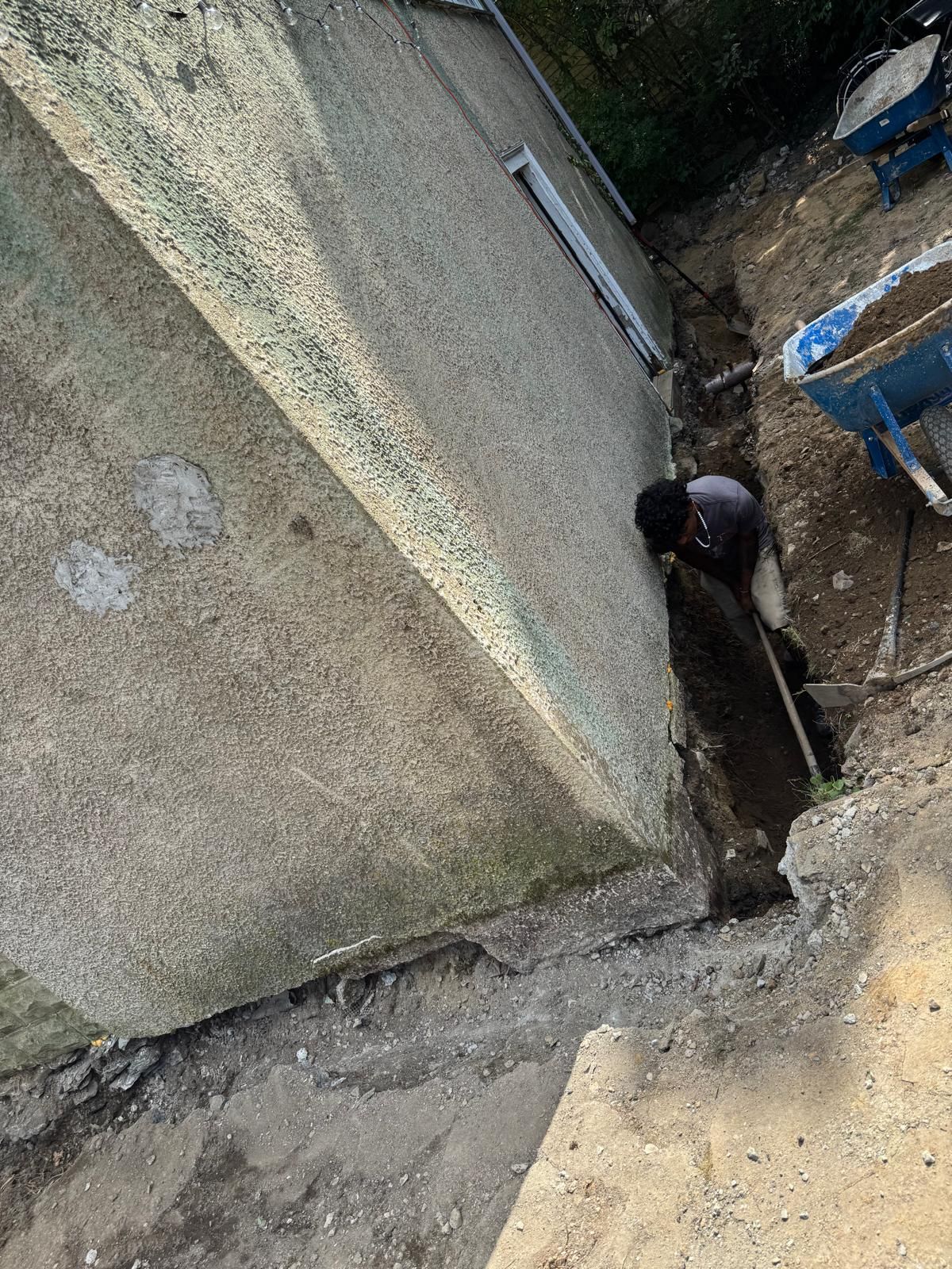 A person digs a trench along the concrete foundation of a building wall next to blue wheelbarrows.